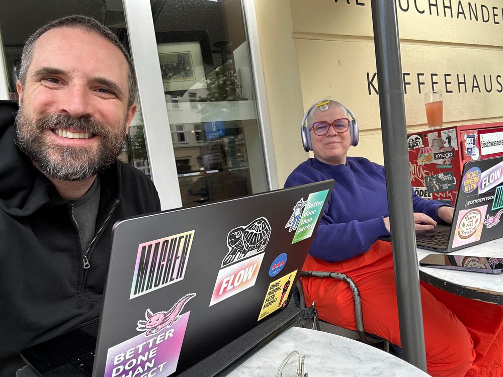Nadja und ein Kollege arbeiten gemeinsam mit Laptops vor einem Café